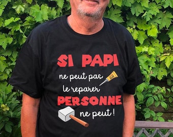 Op de afbeelding: Een man draagt een zwart T-shirt met de tekst "Si Papi ne peut pas le reparer personne ne peut!" in rood en wit. Het shirt heeft ook een cartoon afbeelding van een hamer en een tube lijm.