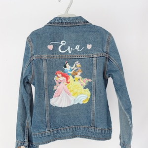 Peut inclure: Une veste en jean bleue avec un nom brodé en blanc "Eva" et des cœurs roses au dos. Le dos présente également un imprimé coloré de princesses Disney : Ariel, Blanche-Neige et Belle.