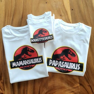 Op de afbeelding: Drie witte T-shirts met korte mouwen en een cirkelvormige afbeelding van een zwart dinosaurus-silhouet op een rode achtergrond. De shirts tonen de tekst MAMANSAURUS, PAPASAURUS en DOUCETTE-SAURUS in witte letters op een zwarte rechthoekige balk.