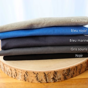 May include: A stack of five t-shirts in different colors, including gray, blue, and black. The t-shirts are folded and stacked on a wooden surface. The colors of the t-shirts are labeled in French: Gris chin&eacute;, Bleu royal, Bleu marine, Gris souris, Noir.
