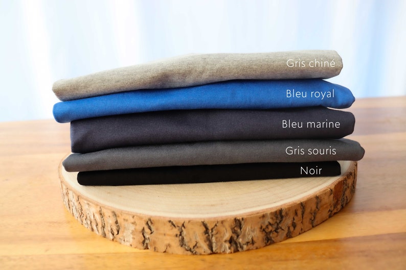 May include: A stack of five t-shirts in different colors, including gray, blue, and black. The t-shirts are folded and stacked on a wooden surface. The colors of the t-shirts are labeled in French: Gris chin&eacute;, Bleu royal, Bleu marine, Gris souris, Noir.