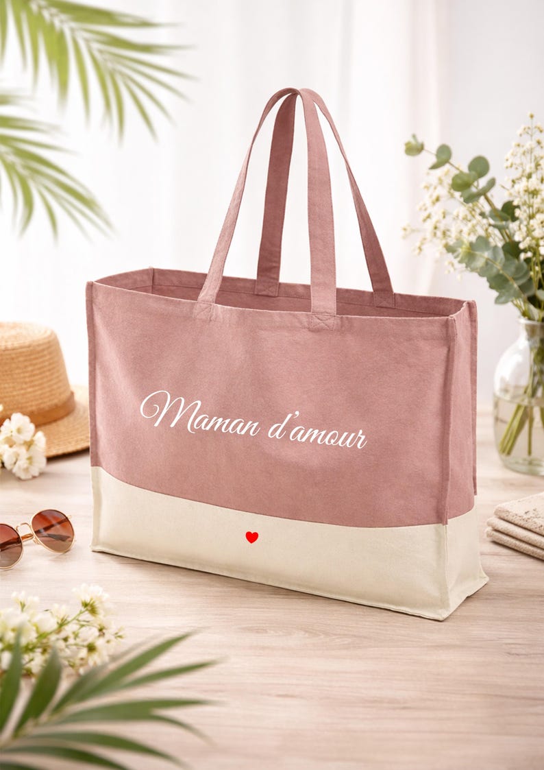 May include: A pink tote bag with a cream-coloured base. The bag features the words "Maman d'amour" in white script and a small red heart. The bag has two handles and is on a wooden surface.