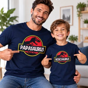 Op de afbeelding: Twee donkerblauwe T-shirts met korte mouwen en een cirkelvormige afbeelding met een rode achtergrond, een zwart dinosaurus-silhouet en een gele rand. Het grotere shirt toont de tekst PAPASAURUS en het kleinere shirt B&Eacute;B&Eacute;SAURUS.