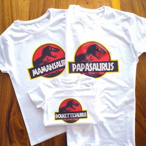 Op de afbeelding: Drie witte T-shirts met korte mouwen en een cirkelvormige afbeelding met een zwart silhouet van een dinosaurus op een rode achtergrond. De tekst MAMANSAURUS, PAPASAURUS en DOUCETTE-SAURUS staat in wit op een zwarte rechthoekige balk.