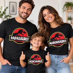 Op de afbeelding: Drie zwarte t-shirts met een cirkelvormig logo met een rode achtergrond en een zwart silhouet van een dinosaurus. De shirts tonen de tekst PAPASAURUS, MAMANSAURUS en B&Eacute;B&Eacute;SAURUS in witte hoofdletters binnen een geel omrand grafisch ontwerp.