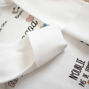 May include: White baby onesie with the text "ma petite vie" and "N'oublie pas de fermer" in black. The onesie has a pink heart and other small colorful illustrations.