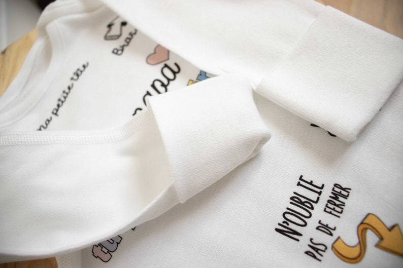 May include: White baby onesie with the text "ma petite vie" and "N'oublie pas de fermer" in black. The onesie has a pink heart and other small colorful illustrations.