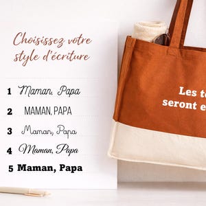 May include: A white sign with French text, beside a brown tote bag. The sign shows font choices for text, including "Maman, Papa." The tote bag has white text that reads "Les textes seront en Blanc."