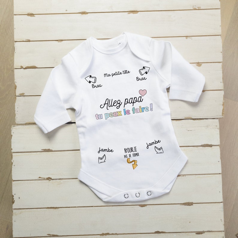 May include: White long-sleeve baby onesie with a pink heart and the text "Allez papa tu peux le faire!" in black. The onesie has arrows pointing to the head, arms, and legs with the words "Ma petite tete", "Bras", "Jambe" in black.