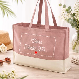 May include: A dusky pink tote bag with a cream base. The bag has two handles and features the text "Votre Texte, LOGO..." inside a dashed white rectangle, with a red heart below. A straw hat and sunglasses are visible.