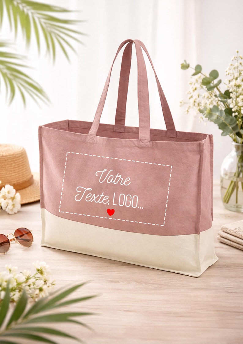 May include: A dusky pink tote bag with a cream base. The bag has two handles and features the text "Votre Texte, LOGO..." inside a dashed white rectangle, with a red heart below. A straw hat and sunglasses are visible.
