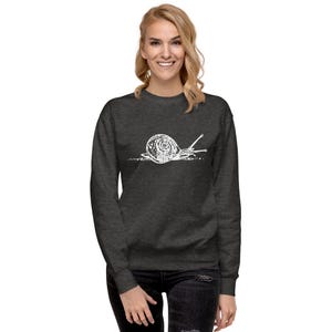 May include: Dark gray sweatshirt featuring a white line drawing of a snail. The snail illustration is centered on the chest. The sweatshirt has long sleeves and a crew neck. The model is wearing black jeans.
