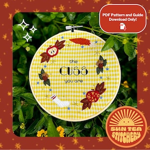 Könnte beinhalten: Ein gelber Gingham-Stoff-Stickrahmen mit dem Text "the CUSS you are". Das Design umfasst gestickte Äpfel, Blätter, Sterne und eine Socke. Das Sun Tea Stitchery-Logo ist sichtbar.