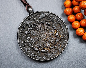 Könnte beinhalten: Ein schwarzer Metallanhänger mit aufwendigen Schnitzereien hängt an einer braunen Schnur. Der Anhänger hat ein kreisförmiges Design mit einem zentralen Symbol, das von kleineren Figuren umgeben ist. Der Anhänger liegt neben einer Reihe orangefarbener Perlen.