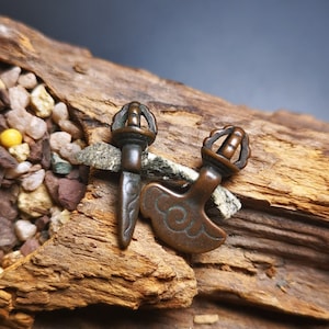 May include: Two small, ornate, bronze-colored metal pendants. One is a stylized sword with a circular handle and the other is a crescent-shaped axe with a circular handle.