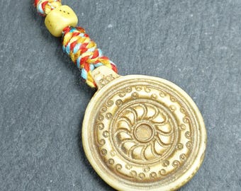 Puede incluir: Un colgante circular beige con un diseño floral en relieve, sujeto a un cordón trenzado multicolor. El cordón presenta hilos rojos, amarillos y azules, con una pequeña cuenta de color claro cerca del colgante.