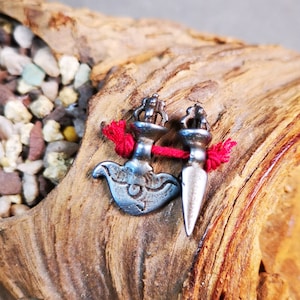 May include: Two silver-toned pendants with red string accents. One pendant is shaped like a hammer with a stylized bird design, and the other is a dagger. The pendants are displayed on a piece of driftwood.