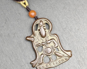 May include: Bronze-colored pendant necklace featuring a stylized figure. The pendant is suspended from a brown cord with gold-colored accents and an orange bead. The pendant has an openwork design with intricate details.
