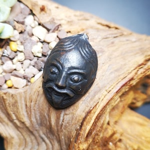 Puede incluir: Un colgante de metal negro con forma de cara con una expresión tallada. El colgante está sobre un trozo de madera a la deriva con suculentas verdes.