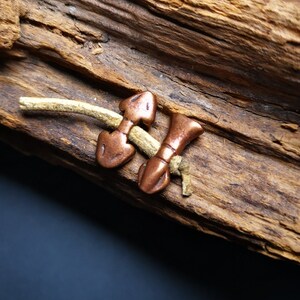 May include: Two copper arrowhead charms on a beige leather cord. The charms are tied together and are lying on a piece of weathered wood.