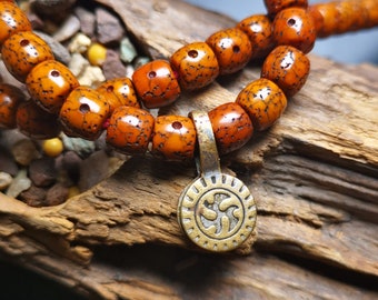 May include: Close-up of a prayer bead necklace with a bronze pendant. The beads are a warm orange-brown color with a textured surface. The pendant has a circular design with an embossed symbol. The necklace is displayed on a piece of wood.