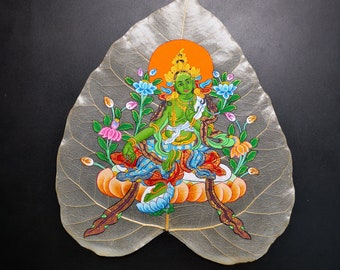 May include: A green deity with a halo and flowers painted on a dried leaf. The deity is sitting in a lotus position with hands in a prayer position. The leaf is a light brown color and has a delicate vein pattern.