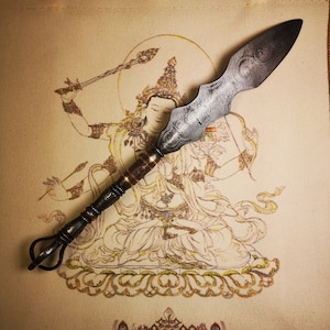 May include: A silver and black metal ritual weapon with a curved blade and a handle with a ring at the end. The weapon is placed on top of a beige fabric with a detailed drawing of a deity holding a similar weapon.