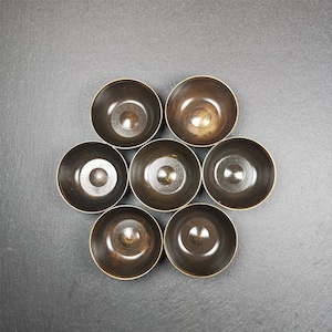 May include: Seven small, round, copper-coloured bowls arranged in a circular pattern on a grey surface.