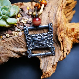 May include: A silver pendant necklace with a detailed, stylized animal face design. The pendant is attached to a brown leather cord with wooden beads.