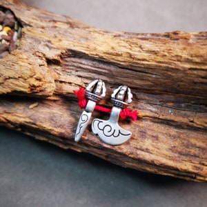 May include: Two silver charms with red string. One charm is a stylized axe with a cloud design. The other charm is a stylized dagger with a crown on top.