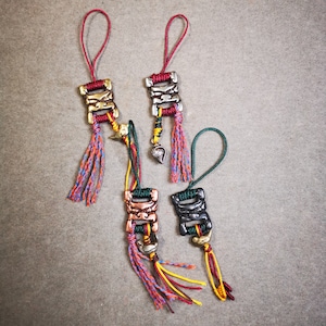May include: Five colorful keychains with intricate metal charms. Each charm is a different color and has a unique design. The keychains are made of braided cords in red, green, yellow, and black.