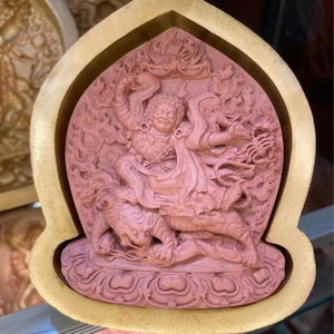 May include: A detailed, pink-toned relief carving of a deity figure, seated atop a tiger-like creature, within a gold-colored frame. The carving features intricate details of the deity's clothing and the creature's fur.