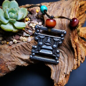 May include: A silver pendant with a detailed design of a face and intricate patterns. The pendant is attached to a brown cord with turquoise and red wooden beads.