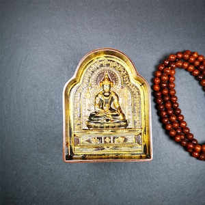 May include: A small, ornate, gold-coloured shrine with a seated Buddha figure inside. The shrine is made of metal and has a dark brown finish. The Buddha is wearing a crown and has a serene expression. The shrine is surrounded by a string of brown wooden beads.