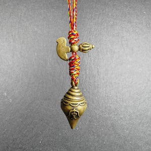 Puede incluir: Un colgante de color bronce con un diseño en forma de pájaro y una base cónica. El colgante está suspendido por un cordón trenzado colorido. El colgante tiene detalles intrincados y es probablemente un artículo decorativo.