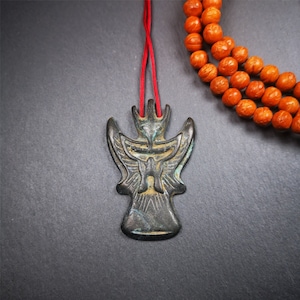 May include: A bronze pendant featuring an intricate design of a bird with outstretched wings. The pendant is suspended from a red cord.