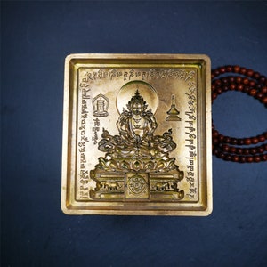 May include: A gold-coloured metal plaque with a raised image of a deity sitting on a lotus throne. The plaque is surrounded by a border of Tibetan script.