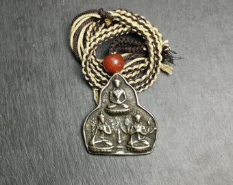 May include: Silver pendant necklace with a three-figure design of seated figures. The pendant is on a brown and cream braided cord with a red bead.