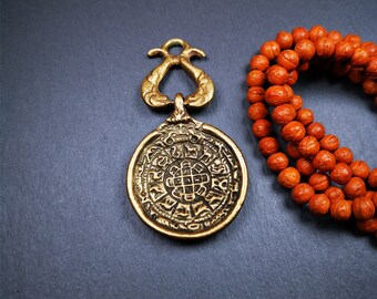 Könnte beinhalten: Antiker Messinganhänger mit aufwendigem kreisförmigem Design und dekorativer Oberseite. Eine Kette aus orangefarbenen Perlen liegt neben dem Anhänger auf einer dunkelgrauen Oberfläche. Der Anhänger hat eine Vintage-Ästhetik.
