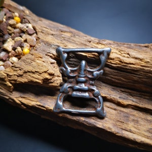 May include: A silver-colored metal bottle opener shaped like a face with a detailed design. The opener is sitting on a piece of weathered wood.