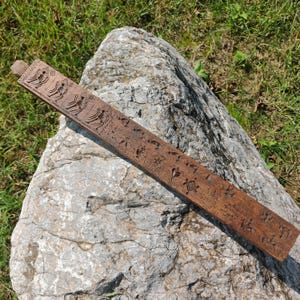 Könnte beinhalten: Ein langer, hölzerner Lineal mit aufwendigen Schnitzereien verschiedener Designs. Der Lineal liegt auf einem großen, grauen Stein.