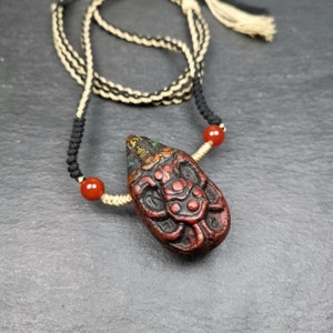 May include: A red, black, and gold pendant with intricate carvings hangs from a black and cream cord with red beads. The pendant is shaped like a teardrop and has a detailed design.