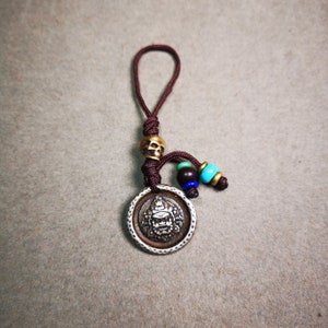 May include: A silver charm featuring an intricate face design, encircled by a circular border. The charm is attached to a brown cord with a skull bead and colourful beads.