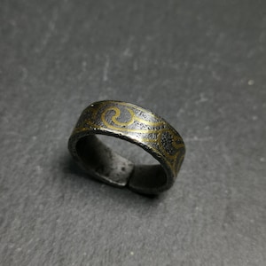 Può includere: Un anello argentato con finitura scura e strutturata. L'anello presenta un motivo a spirale dorata ripetuto attorno alla fascia. L'anello è mostrato su una superficie grigio scuro.