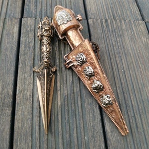 May include: A pair of ornate, copper-colored daggers with intricate designs. One dagger is in a sheath with a decorative design of four faces. The other dagger has a handle shaped like a hand with a face.