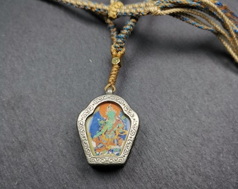 May include: A silver pendant necklace with a small, framed image of a green deity. The pendant is attached to a braided cord with brown and blue threads.