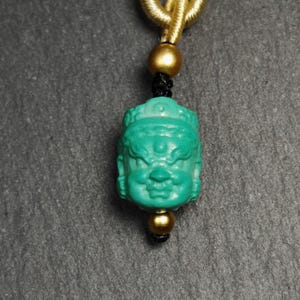 May include: A turquoise green pendant with a carved face, hanging from a gold and black cord.