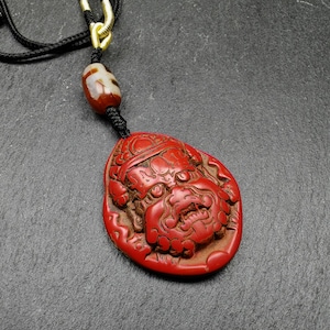 May include: A red carved pendant with a detailed face and a black cord necklace. The pendant is attached to the cord with a brown and white bead.