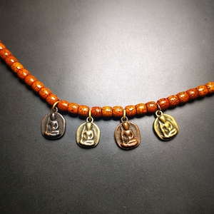 May include: A necklace made of orange wooden beads with four small metal charms depicting seated figures. The charms are in different shades of gold and brown.