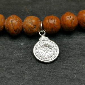 Puede incluir: Un brazalete de cuentas de madera marrón con un dije de plata con diseños intrincados. El dije es redondo y tiene un centro elevado.